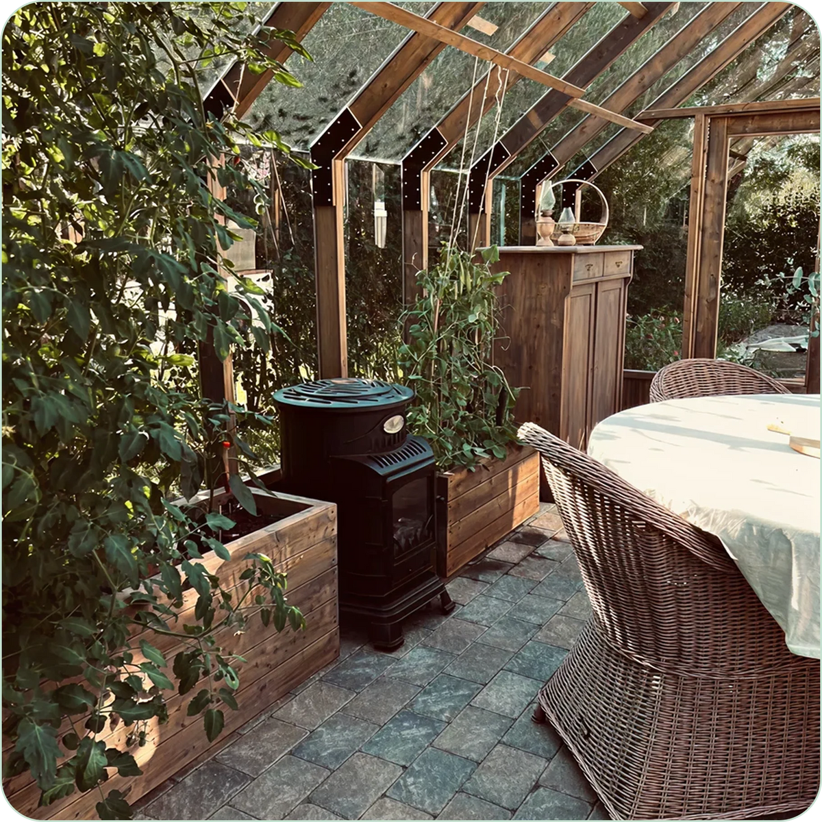 Interior of a wooden Nordwood greenhouse with plants, a stove, and wicker chairs